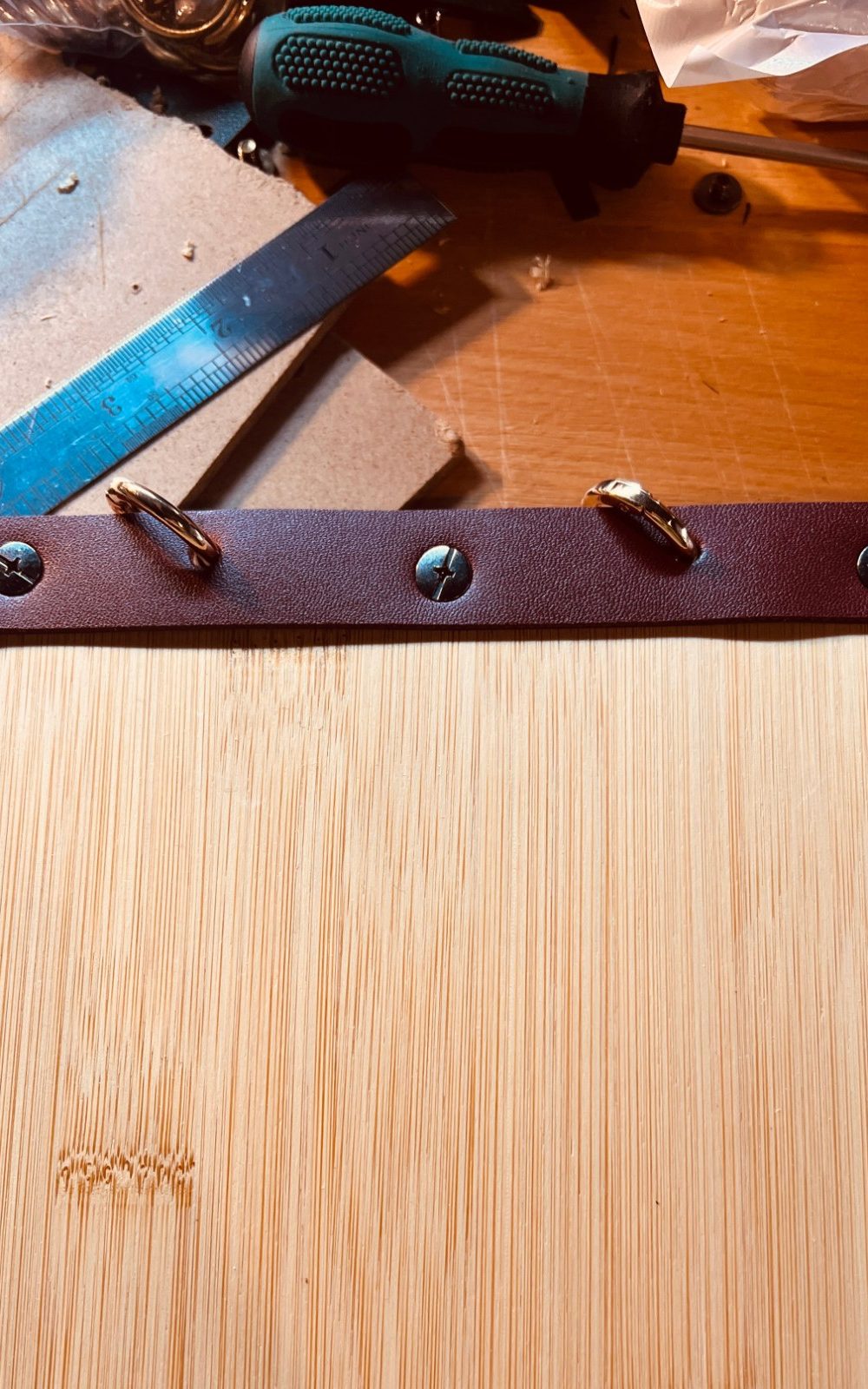 A bamboo menu board with a burgundy cuff and two rings on a workbench, next to a steel ruler and screwdriver